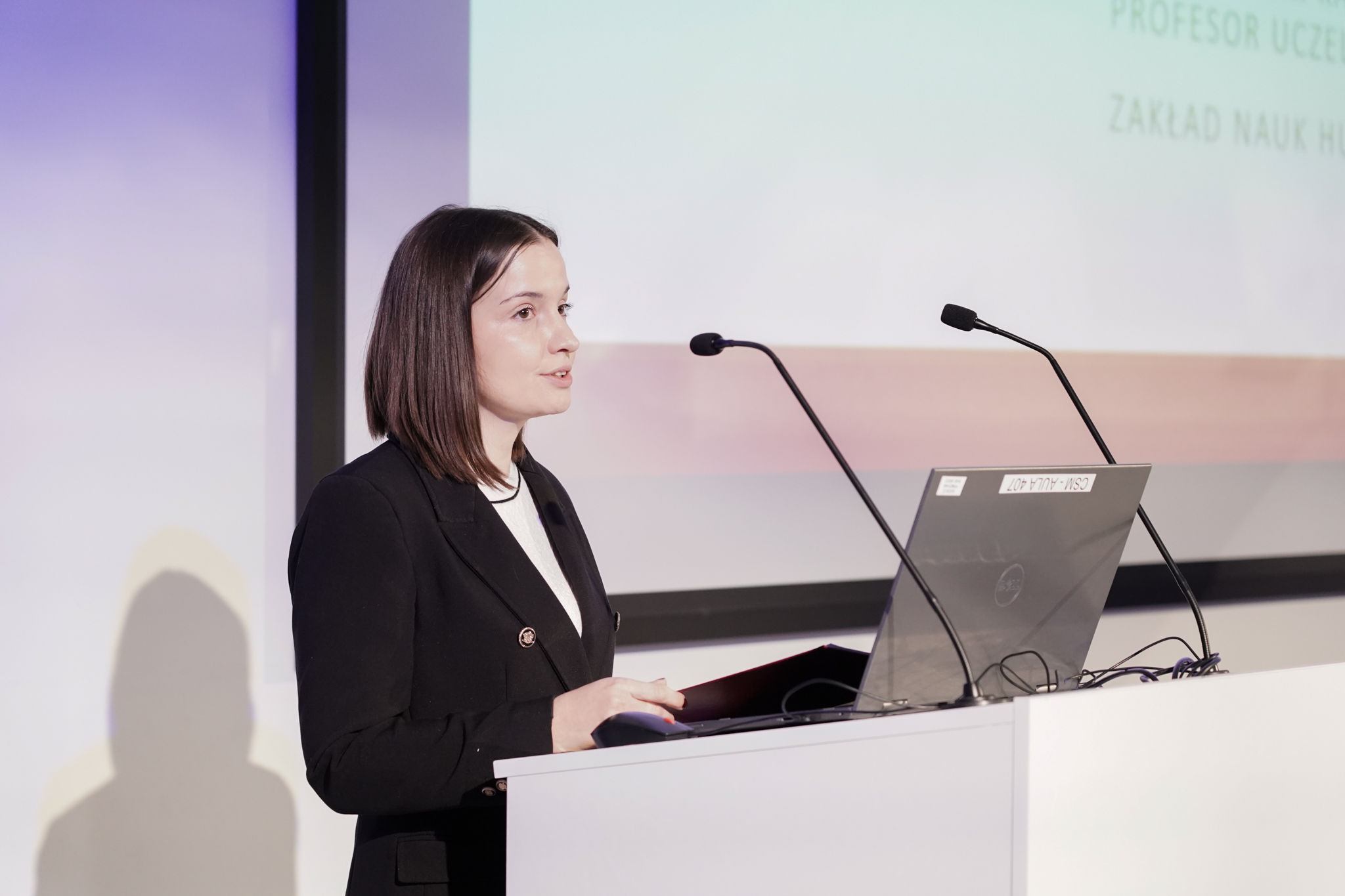 Elegancko ubrana młoda kobieta za m&oacute;wnicą z dwoma mikrofonami, z tyłu wyświetla się część prezentacji.